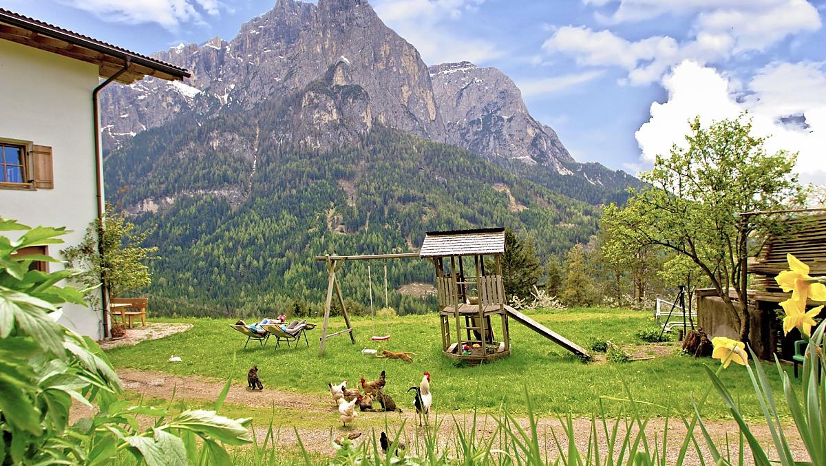 Il nostro maso si trova ai piedi dell'Alpe di Siusi, sullo sfondo lo Sciliar ancora innevato, in primo piano il parco giochi e il prato per prendere il sole.
