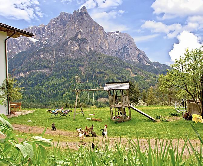 Il nostro maso si trova ai piedi dell'Alpe di Siusi, sullo sfondo lo Sciliar ancora innevato, in primo piano il parco giochi e il prato per prendere il sole.
