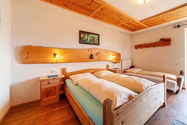 Schlafzimmer mit Doppelbett und Einzelbett, Holzboden und Möbeln aus Zirbenholz. Zugang zum großen, überdachten Balkon.