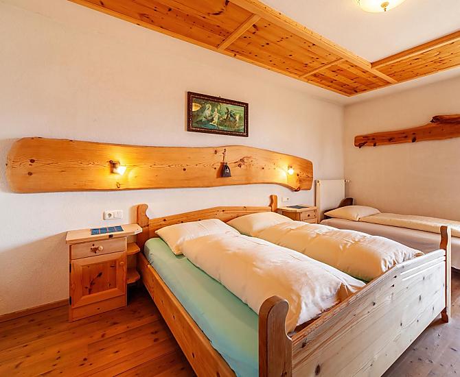Schlafzimmer mit Doppelbett und Einzelbett, Holzboden und Möbeln aus Zirbenholz. Zugang zum großen, überdachten Balkon.